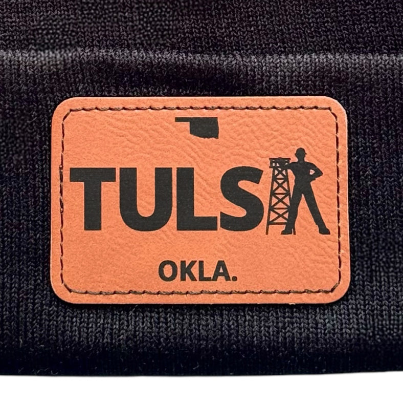 Close-up of a black knit beanie with a detailed leather patch of the Tulsa Driller and Oklahoma emblem.