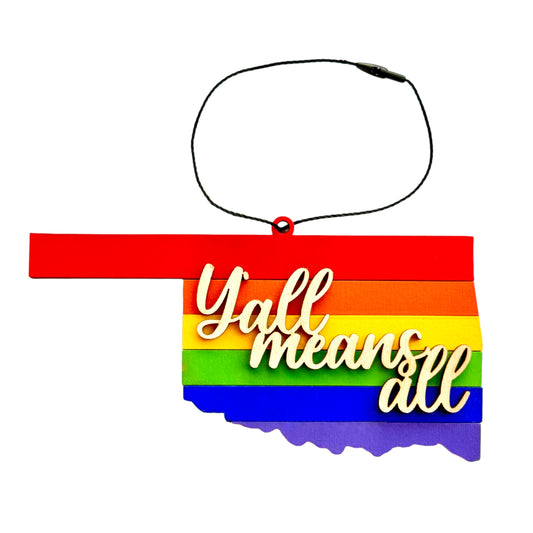 A colorful wooden ornament shaped like Oklahoma with vibrant rainbow stripes and the words “Y’all Means All” in bold script.